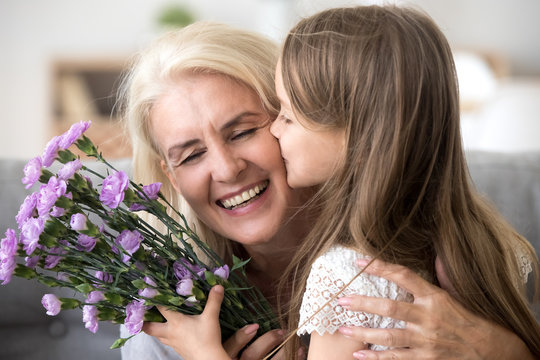 Little Preschool Granddaughter Kissing Happy Older Grandma On Cheek Giving Violet Flowers Bouquet Congratulating Smiling Senior Grandmother With Birthday, Celebrating Mothers Day Or 8 March Concept