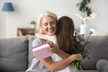 Happy senior grandma hugging granddaughter thanking for present holding flower bouquet, smiling...
