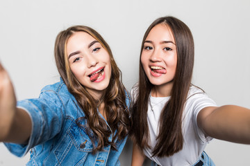 Portrait of two joyful multiracial women taking selfie on smartphone, with tongue out isolated over gray background