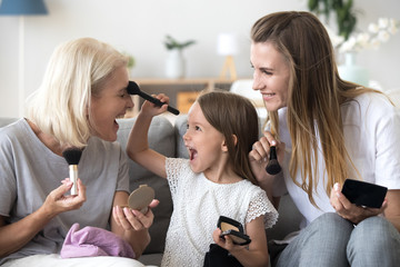 Obraz na płótnie Canvas Excited little child girl holding brush puts powder on grandmas face doing make-up with mom and grandmother, happy kid granddaughter applying makeup on granny having fun in 3 generations women family
