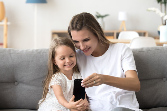 Happy smiling mother and cute preschool daughter laughing taking selfie on smartphone, cheerful mom and little child girl playing making photo on mobile, mommy and kid having fun with phone at home