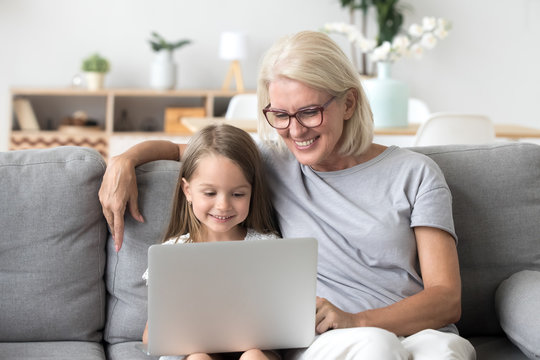Smiling senior grandma and cute little kid granddaughter watching cartoons on laptop together, happy older grandmother playing game or shopping ordering buying online with child girl on computer