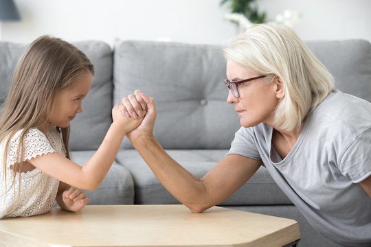 Serious old grandma and little granddaughter arm wrestling, grandchild girl and grandmother competing playing together, grandparents and children generations confrontation, family conflicts concept