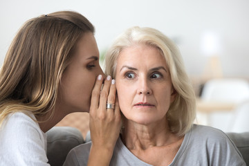 Shocked older woman listening to young female whispering in ear, friend gossiper telling secret...