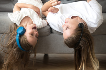 Excited carefree mom and little child in headphones enjoy listening to music together, smiling kid daughter and happy mother lying upside down on sofa laughing having fun hearing songs in earphones