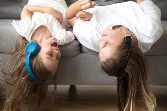 Excited Carefree Mom And Little Child In Headphones Enjoy Listening To Music Together, Smiling Kid Daughter And Happy Mother Lying Upside Down On Sofa Laughing Having Fun Hearing Songs In Earphones