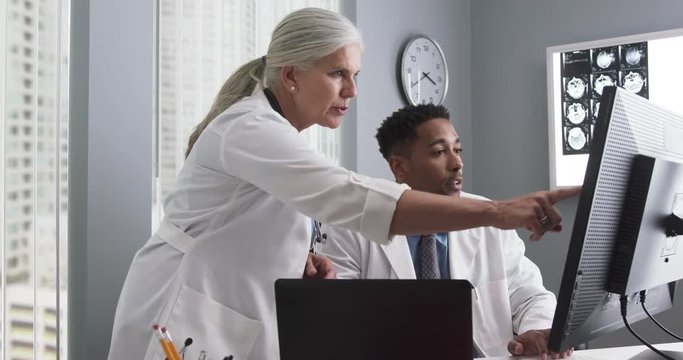 Portrait of millennial black doctor using computer while senior colleague directs him. Two doctors working inside medical office looking at computer monitor and typing