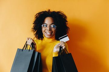 Black friday. Shopping. Afro American girl in eyeglasses is holding shopping bags and a credit card and smiling, on a yellow background
