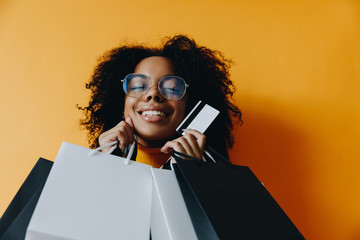 Black friday. Shopping. Afro American girl in eyeglasses is holding shopping bags and a credit card and smiling, on a yellow background