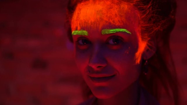 A beautiful young sexy girl with glowing paint on her face and hair in black light talking. Pretty woman with glowing bodyart in black lamp light.