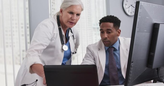 Female caucasian senior doctor working with young african-american colleague on laptop computer. Close up of two medical professionals working together with technology inside office