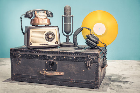 Retro radio from 60s, old copper telephone, studio microphone from 50s, and gold colored vinyl disk record circa 70s, headphones on aged classic travel trunk. Vintage style filtered photo
