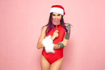Girl with purple hair tips and tattoo on her arm dressed in red swimsuit, Santa's hat and white fur bracelets holding a lollipop in front of her face on the background of pink wall