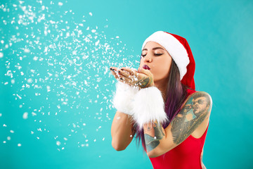 Bright girl with tattoo on her arm dressed in red swimsuit, Santa's hat and white fur bracelets blows snowflakes from your palms on the background of blue wall