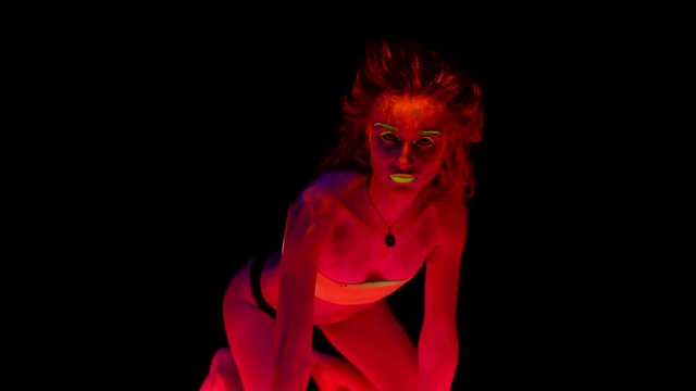 A beautiful young sexy half-naked standing kneeling on all fours with glowing paint on her body in black light looking provocative. Pretty woman with glowing bodyart in black lamp light.