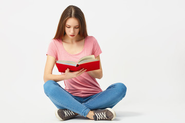 Beautiful girl dressed in a pink t-shirt, jeans and sneakers sits on the floor and reads a book on a white background in the studio