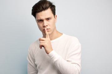 Dark-haired guy dressed in a white long sleeve t-shirt keeps his finger on his lips on a white background