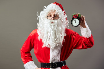 Santa Claus in glasses holds clock a clock that shows five to twelve in his hand on the gray background
