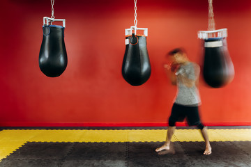 Guy dressed in the grey t-shirt and black shorts works out with a black punching bag against a red wall in the gym