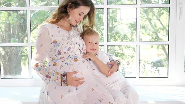 Little boy kissing pregnant mother in belly sitting together on window sill in sunlight, slow motion