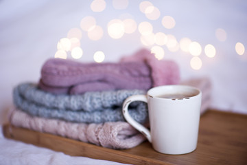 Fototapeta na wymiar Cup of coffee with stack of knitted clothes over Christmas lights. Winter season.