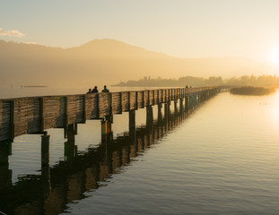 long wooden pier and boardwalk over Lake Zurich near rapperswill in golden evening light with silhouette of pedestrians and people walking and mountains in the background