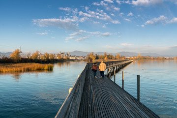 landscape view of marsh and lake shore with the town of Rapperswil in evening light and a long wooden boardwalk in the foreground and a couple going for a leisurely walk