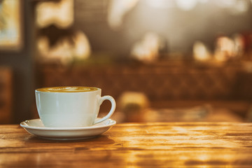 Cappuccino coffee in a white cup on a wooden table with a modern coffeeshop in the background, selective focus.