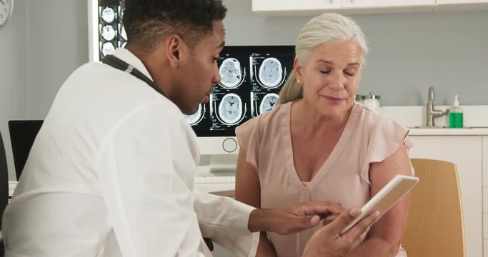 Medical doctor and senior woman looking at tablet computer indoors health clinic. Young black doctor using electronic pad to show female patient test results from her ct scan