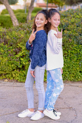 Close up of a two cheery stylish girls showing peace and rock and roll gesture over nature autumn summer background. Two young happy hipster kid friends with ponytail standing together and having fun.