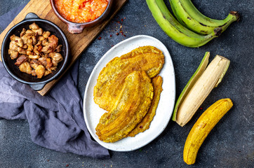 COLOMBIAN CARIBBEAN CENTRAL AMERICAN FOOD. Patacon or toston, fried and flattened whole green plantain banana on white plate with tomato sauce and chicharron Black background, top view