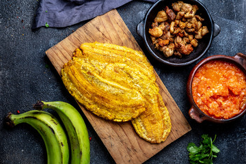 COLOMBIAN CARIBBEAN CENTRAL AMERICAN FOOD. Patacon or toston, fried and flattened whole green plantain banana on white plate with tomato sauce and chicharron Black background, top view