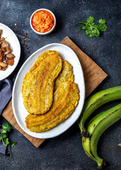 COLOMBIAN CARIBBEAN CENTRAL AMERICAN FOOD. Patacon or toston, fried and flattened whole green plantain banana on white plate with tomato sauce and chicharron Black background, top view