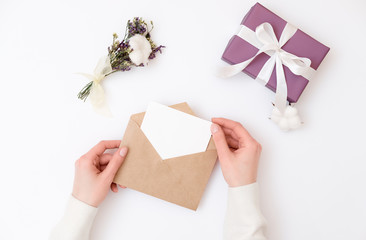 Woman's hands holding kraft envelope with blank wedding invitation card, Gift box, floral minimalist bouquet laying on woman desk. Creative mock up layout on white background, greeting card, top view.