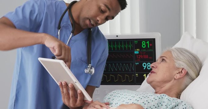 Male registered nurse using tablet to show senior woman test results from her surgery. Elderly female patient lying in bed looking at electronic pad with nurse