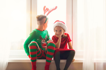 Brother and sister sitting restless on window sill at christmas time, looking out the window,...