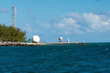 Turquoise ocean laps a rocky pier topped by white radar domes and palm trees beneath a bright blue sky—coastal facility and scenic shoreline. © Daniel Wedeking