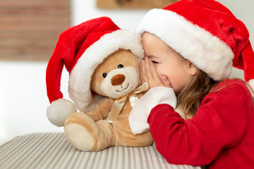 Cute young girl wearing santa hat whispering a secret to her teddy bear christmas present toy. Cheeky kid sharing secrets with teddy bear.