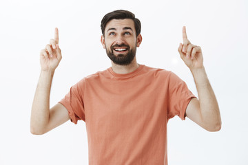 Fototapeta premium Happy curious attractive european man with dark beard in casual t-shirt raising index fingers looking and pointing up wondered and delighted enjoying nice view on stars over gray background