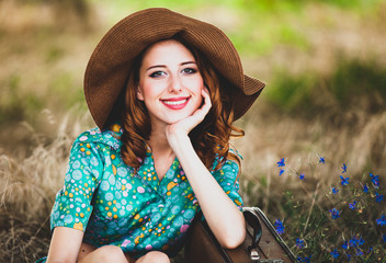 Young redhead girl in hat and dress with suitcase sitting at autumn season grass.