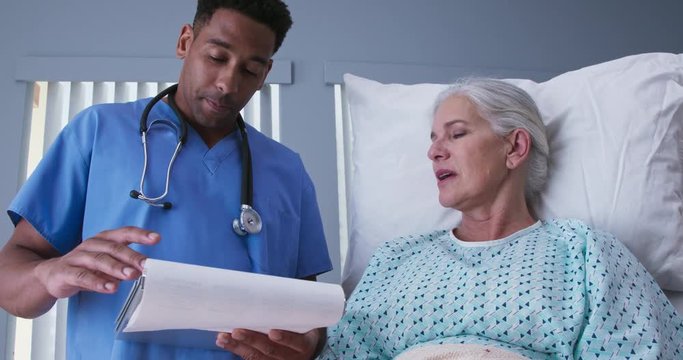 Millennial male nurse reading doctors notes to elderly female patient. Ill senior woman resting in hospital bed while talking to young male RN
