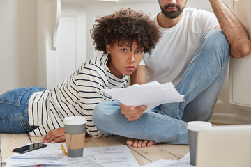 Business multiethnic female and male colleagues think over financial report, pose on floor in modern apartment, drink hot coffee, work with paper documents, use laptop and high speed internet