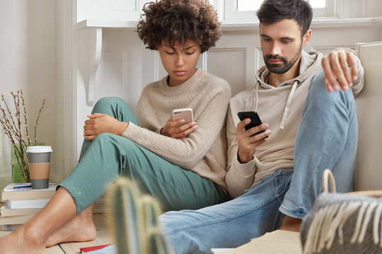 Image of couple in love sit at living room on floor together, use cell phones for surfing online, connected to wifi at home, spend leisure time with modern technologies, pile of books with coffee near