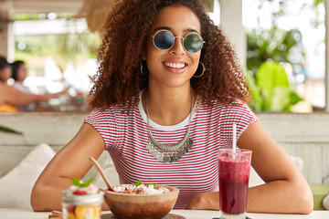 Young woman in fashionable round shades has fresh drink and eats tasty exotic dish, smiles broadly, rests at cafeteria, dressed in t shirt, enjoys good time. People, eating and rest time concept