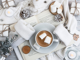 A cup of coffee with marshmallows stands on the open book. A warm sweater. New Year's and Christmas. Christmas tree and gifts of jewelry, bracelets with charms. Huggy concept.