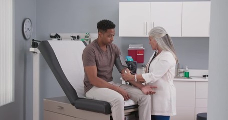 Mature female medical practitioner measuring blood pressure with monitor and stethoscope of a young male patient. Attractive black man having his blood pressure measured by senior white doctor - Powered by Adobe