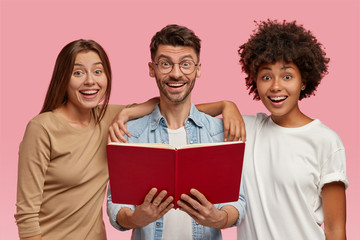 Indoor shot of cheerful handsome man and his two sisters embrace and stand closely, read book, have...