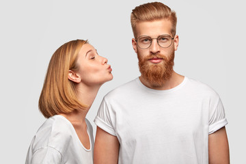 Beautiful European woman with bobbed hairstyle, dressed in casual clothes, stands sideways near her boyfriend, going to kiss him in cheek, enjoy togetherness, isolated over white wall. Relationship