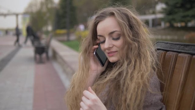 Close up beautiful young blonde woman with curly hair in trendy coat phone coversation sitting in the city park on bench on a warm day looking at the camera and smiling