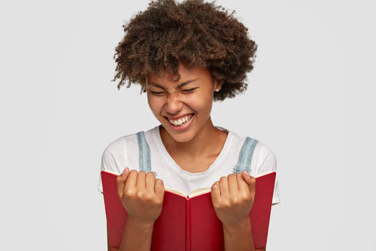 Joyful woman laughs happily while reads funny story from book, shows white teeth, squints face as smiles, dressed in casual outfit, isolated over white background. People, hobby and reading concept